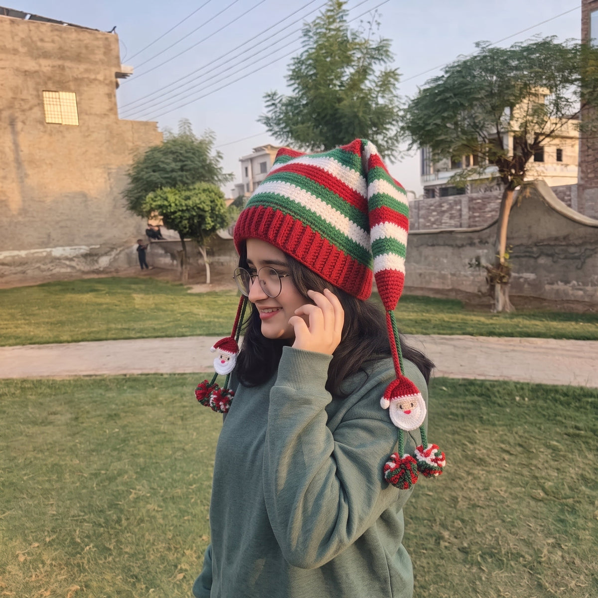 Woman holding mug wearing crochet Christmas elf hat with Santa face pom-poms in nature setting